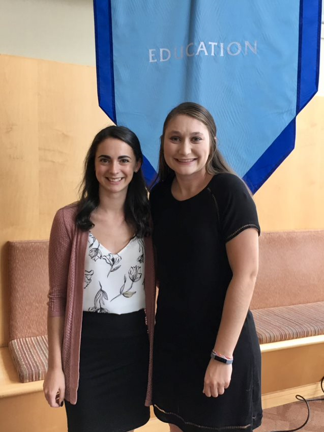 two young female students in professional dress stand side by side with a blue banner behind them that says Education