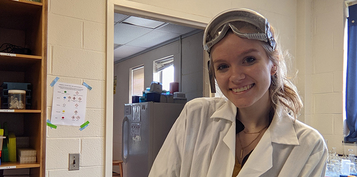 Courtney Smith is wearing a white lab coat and standing in a lab. She is smiling and looking at the camera.