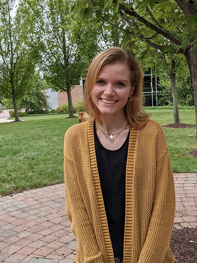 Courtney Smith smiling at the camera wearing a yellow cardigan, standing outside.