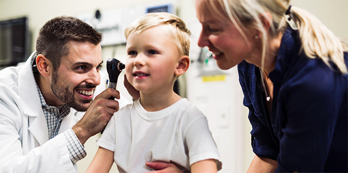 A man in a white coat leans down and examines a child's ear as he sits on a table in a doctor's office. A woman stands on the other side of him, smiling.