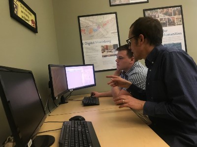 A professor sits at an office computer with a student.