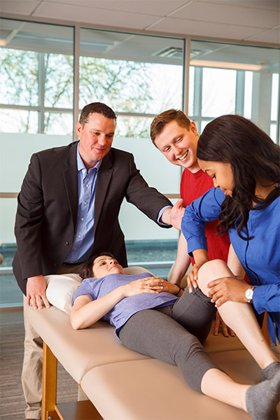 Two men and a woman stand around a patient as she lays on a bed with her knee up, getting pushed on by the woman.