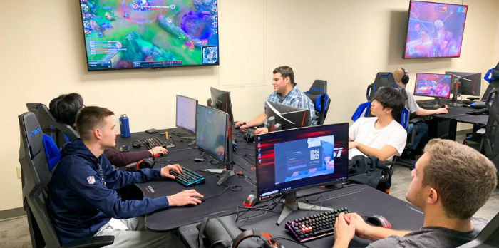 Male students sit at computers while looking up at a monitor while playing competitive video games.