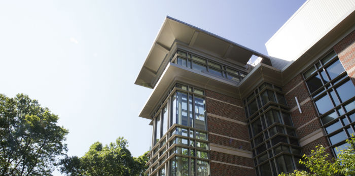 A brick building with many glass windows is shown in front of a blue sunny sky. Green trees sit around the building.