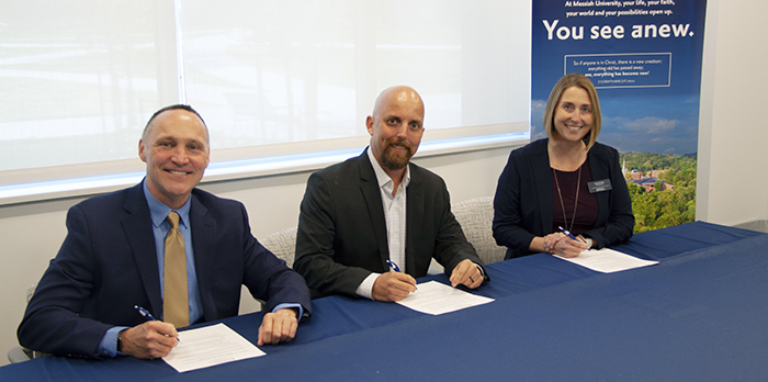 Three individuals, 2 men and 1 woman, sit at a table with a blue tablecloth. They are signing a contract and smiling at the camera.