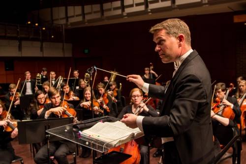 A male conductor surrounded by string instrument players.