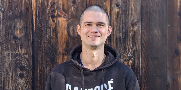 A man with a shaved head and black hoodie stands in front of a wooden background smiling.