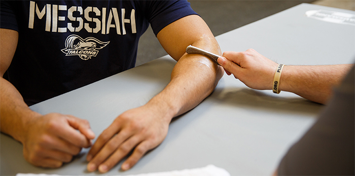A man wearing a Messiah t-shirt has his arms resting on a table. Another man holds a device to his forearm.