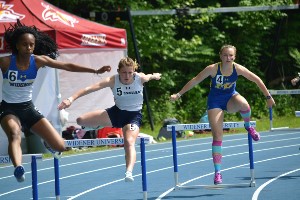 Three female runners jump hurdles on a track.