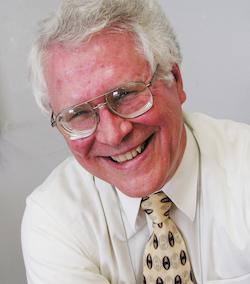 Picture of a white man with white-gray hair smiling. He is wearing a yellow tie and a white dress shirt.