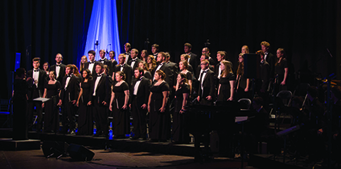 A choir of men and women stands on risers in black outfits in front of curtains.