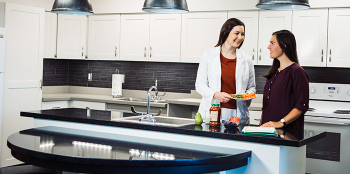 Two women stand together in a kitchen and look at each other. There is fruit and a jar on the counter in front of them. The one on the left holds a plate of veggies and wears a white lab coat.