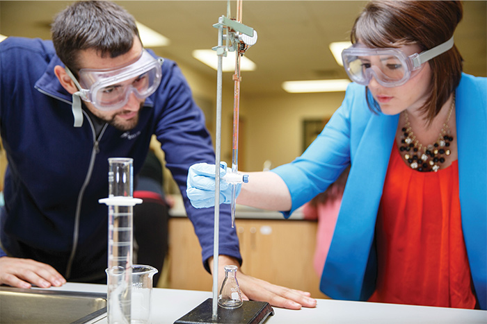 A man and a women wear safety goggles and lean over a counter as the woman holds something up to a test tube. A flask and a beaker sit on the counter as well.