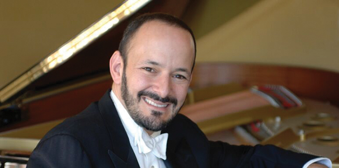 A man sits smiling in front of a grand piano with his arm on it.