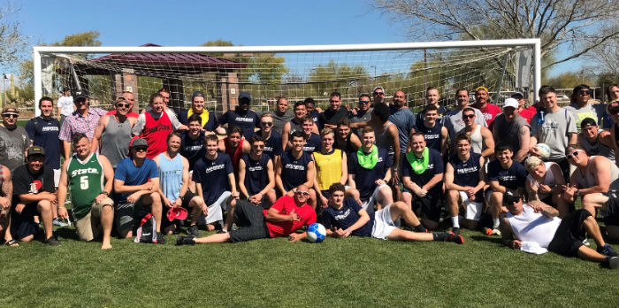 A large group of male soccer players, some older and some younger, pose for a group photo in front of a soccer net on green grass.