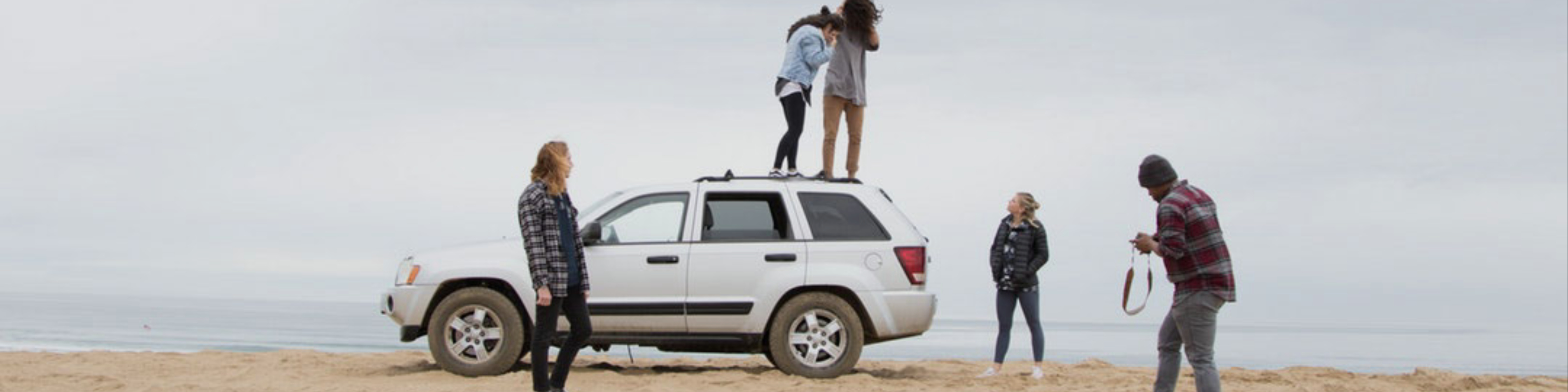 A group of young adults around a car. Two are standing on top of the car, one is holding a camera and the rest are standing around.