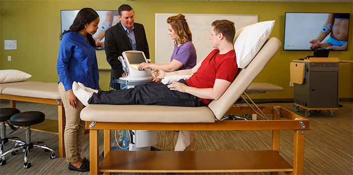 A man lays on a table while a professor and two students stand around him, pointing at a screen.