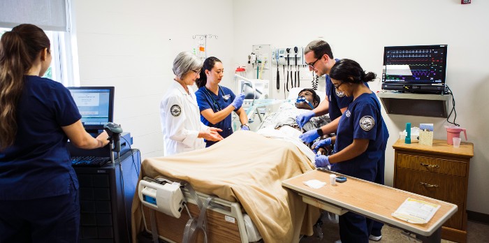 Nursing students and a professor work on a dummy laying in a hospital bed.