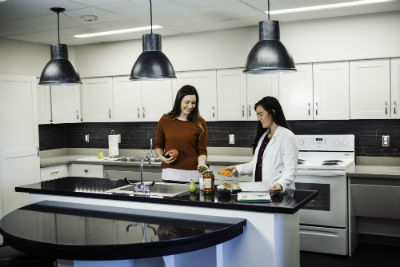 Two females stand at a kitchen counter smiling and making food. One female is wearing a lab coat.