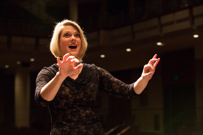 A woman with her mouth open and her hands up as if she is conducting. She is in some sort of hall and is facing the camera, but isn't looking directly at it.