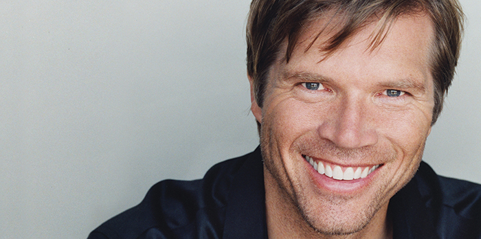 A man smiles in front of a plain grey background.