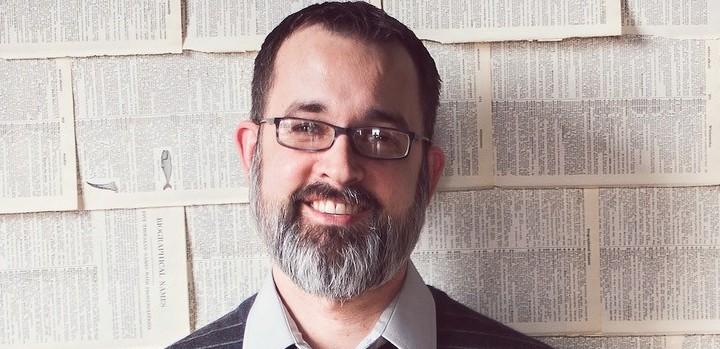 A man with glasses standing in front of a cinderblock wall and smiling.