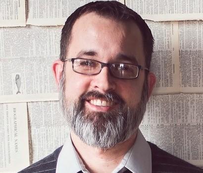 A man with glasses standing in front of a cinderblock wall smiling.