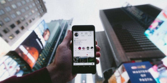 A hand holds an iPhone up toward the sky with city skyscrapers in the background.