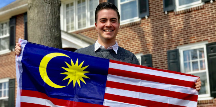 A male student stands in front of a brick house smiling and holding the flag of Malaysia.