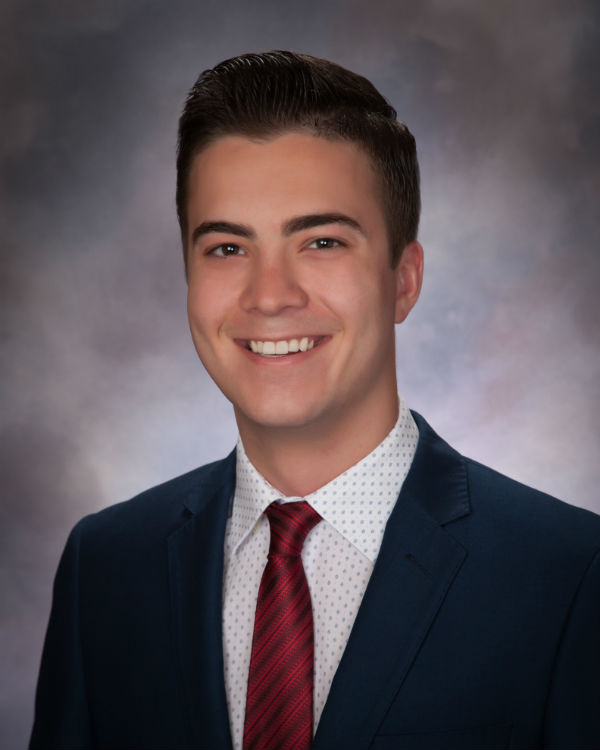 A male student smiles for a headshot wearing a suit and tie with a plain background.