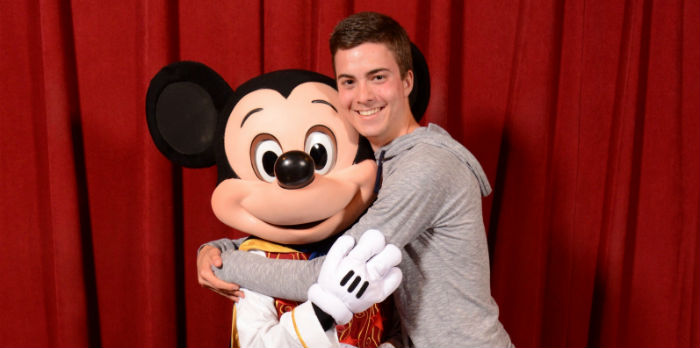 A male Caucasian student hugs Mickey Mouse in front of a red curtain.