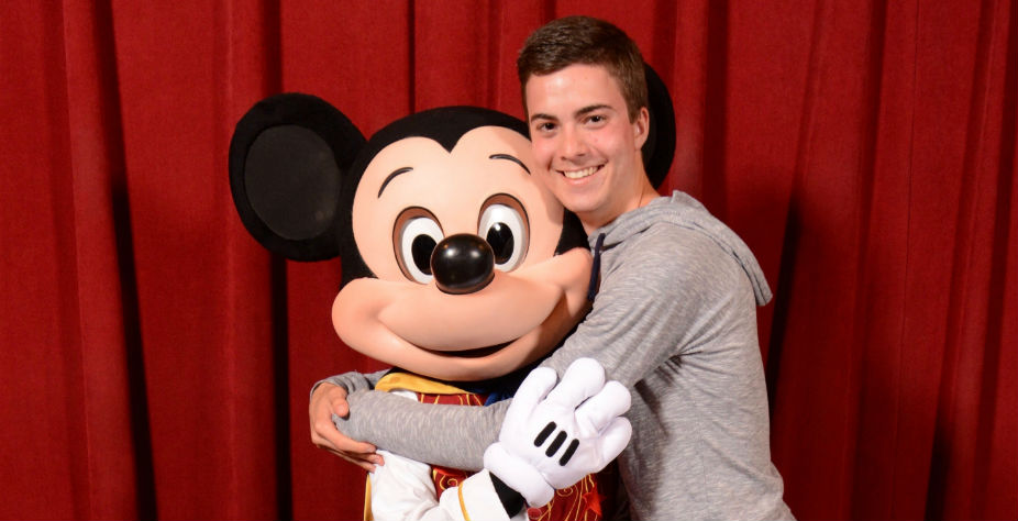 A male student hugs Mickey Mouse in front of a red curtain.