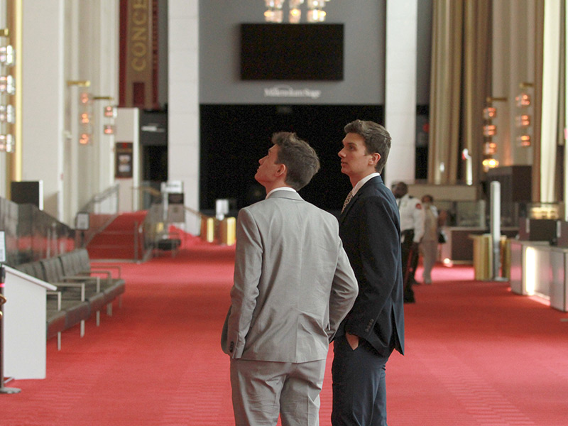 Two male students wearing suits stands inside a room with red carpet.