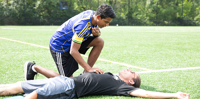 A man kneels over another man who is laying on his back in a soccer field, arms outstretched.