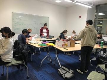 A group of students standing and sitting in a white room. Some are working on a project that involves paper.