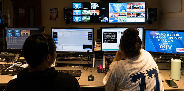 Two students sit in front of six screens and direct a live broadcast.