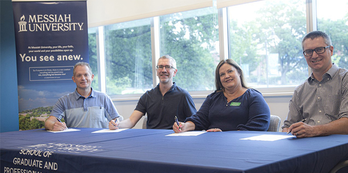 Three men and one woman sit at a table and smile for the camera. They each hold a pen in their hand as they sign a piece of paper. Behind them is a Messiah banner.