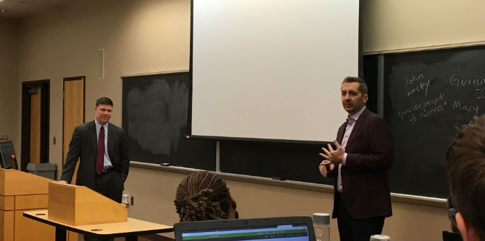 Two men stand in the front of a classroom in business clothes in front of a chalkboard as one talks to students.