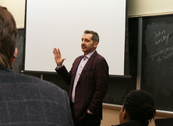 A man stands in front of a classroom wearing a suit jacket and speaking to the class.