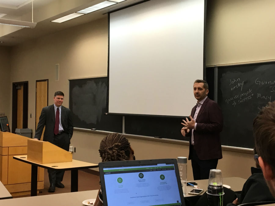Two men in business clothing stand in the front of a classroom as one speaks to students.