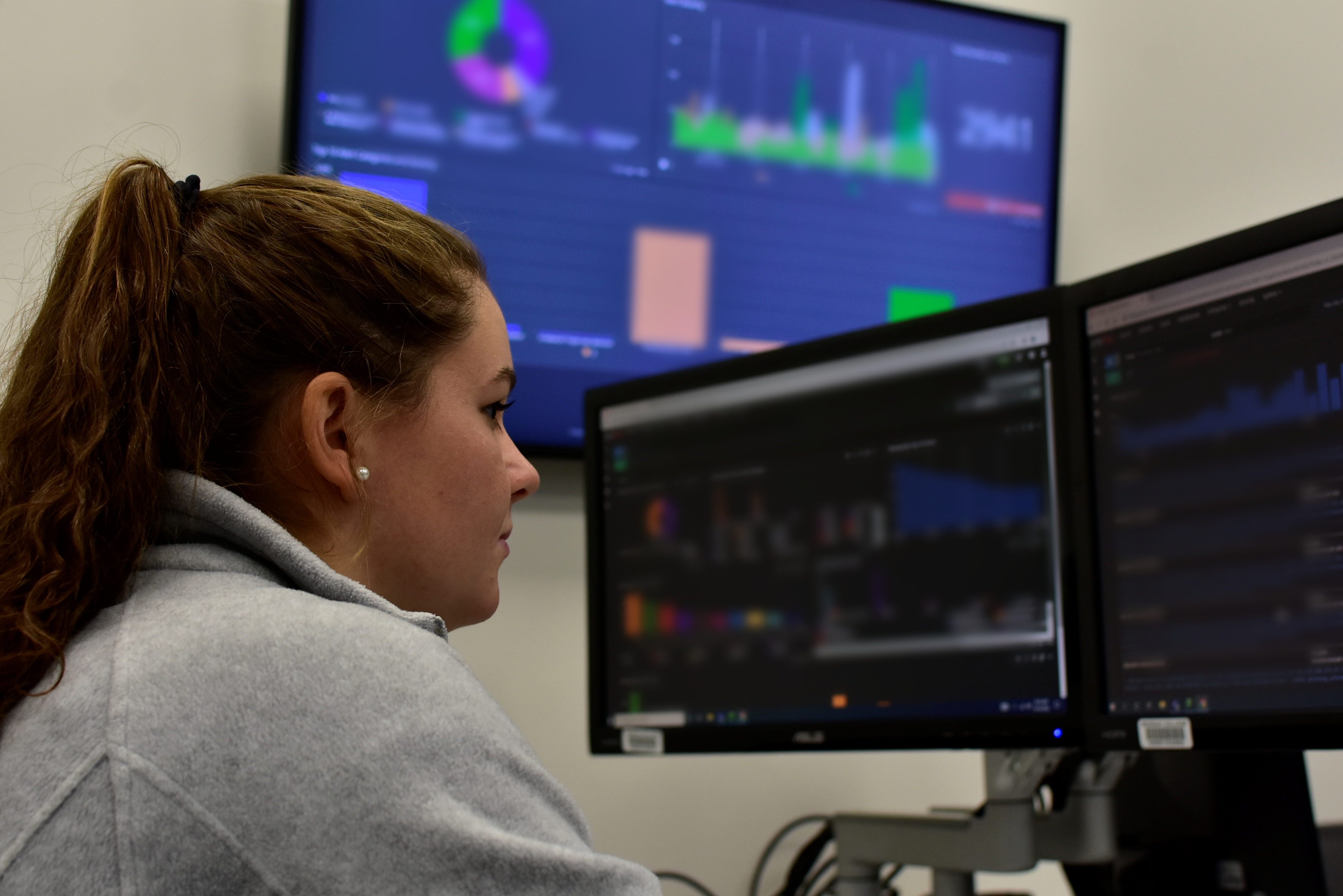 Woman sits facing two computer screens. You can see data and graphics on the screen.