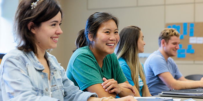 Two female students smile and sit at a table in a classroom.