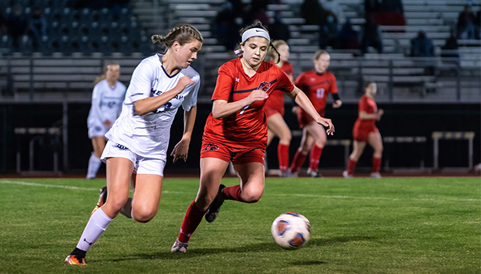 Two female soccer players race across the field for the ball. One wears white and one wears red.