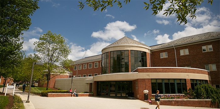 Leaves frame a brick building against a blue sky. A student walks in front of the building.