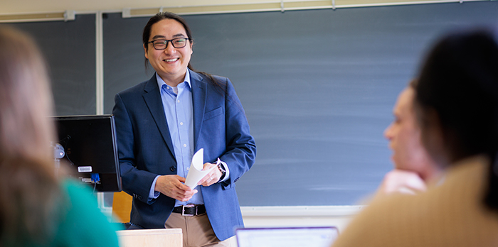 Jason Renn stands in front of a classroom, wearing a navy blazer, and smiles at two students as he teaches.