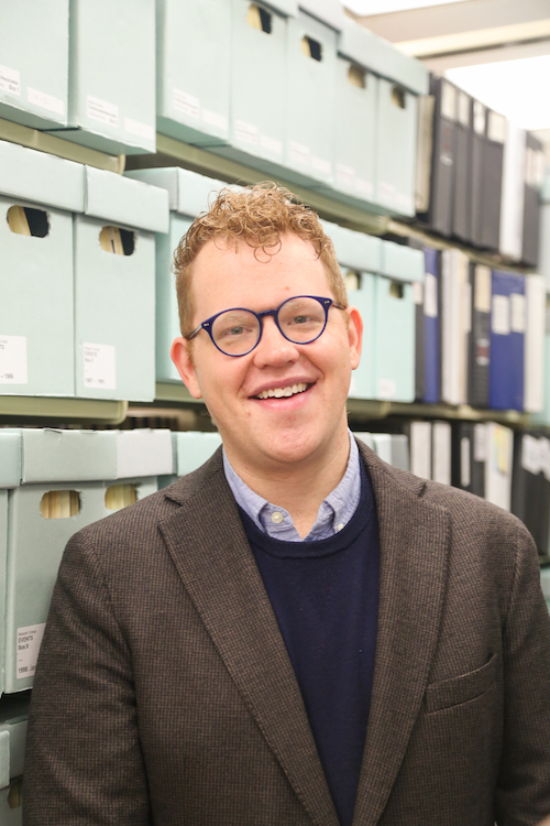 A man with glasses wearing a blue sweater and brown blazer stands in front of a shelf of blue boxes