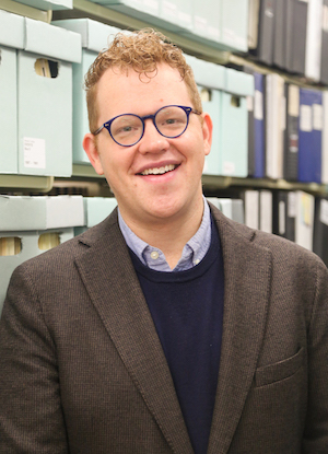 A white man with glasses wearing a blue sweater and a brown blazer standing in front of a shelf of blue boxes