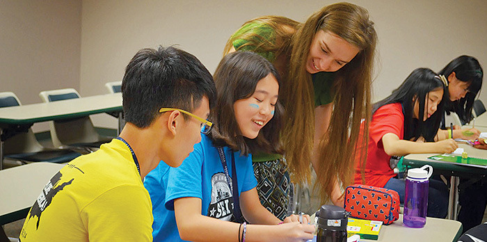 A female teacher bends down to help two students sitting at a table.