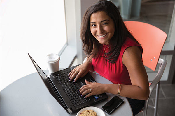 A woman in a red shirt sits at a table and types on her laptop. She looks up at the camera and smiles.