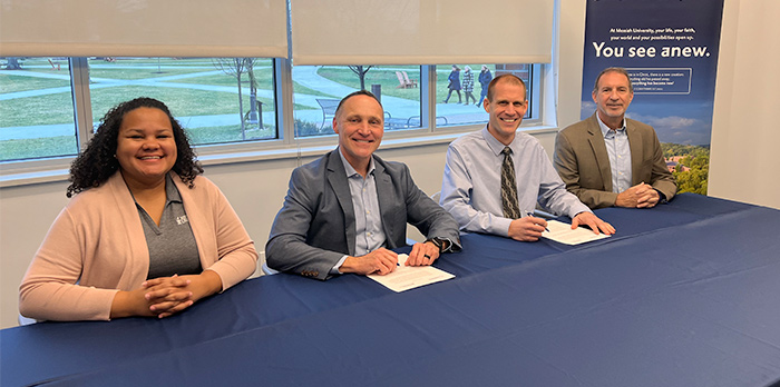 One woman and three men sit at a table with a blue tablecloth, their hands folded in front of them.